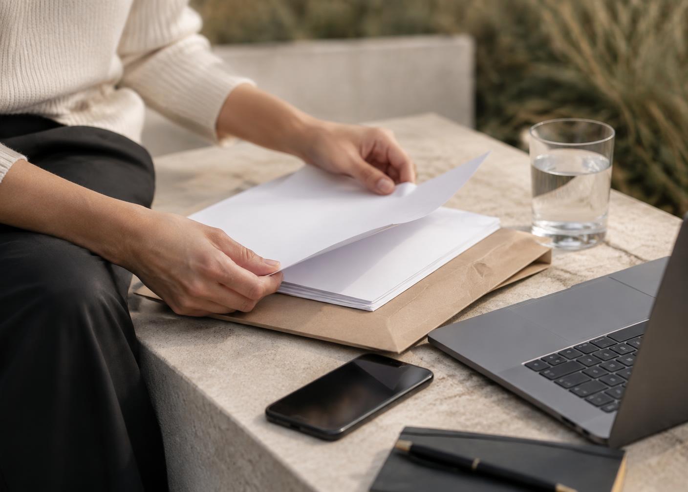 Hands sorting papers into a folder beside a laptop on a stone bench